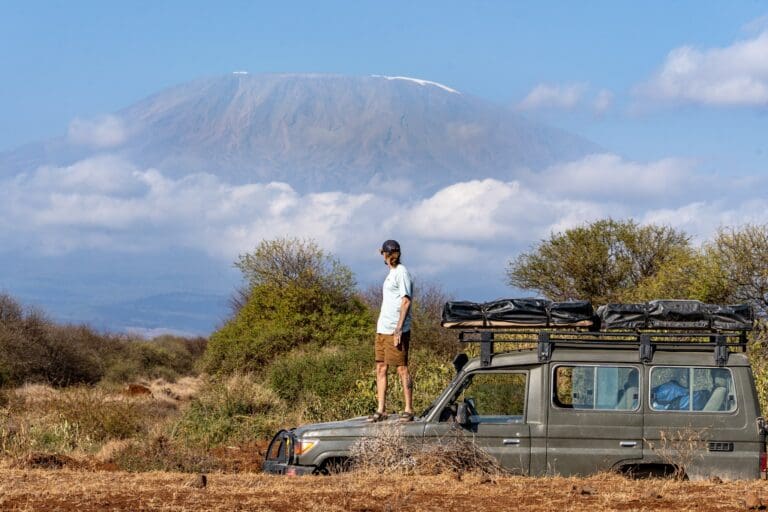 Mann steht auf einem Safarifahrzeug, im Hintergrund der Mount Kilimanjaro